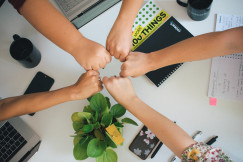 The photo presents five people bringing their fists together over a desk with laptops, notebooks, mugs and a potted plant.
