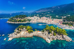 The photo presents an aerial view of a coastal town and harbour, with rocky, tree-covered islets in turquoise water and forested mountains behind.
