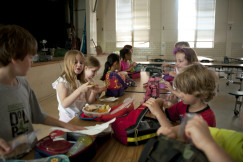 The photo presents children unpacking and eating their lunches together at a long table in a school cafeteria, with colourful lunch bags and food containers.