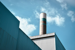 The photo presents an industrial chimney rising above factory buildings under a blue sky.