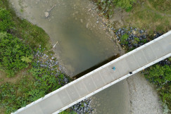 The photo presents an overhead view of two people on a footbridge crossing a shallow, rocky river surrounded by green vegetation.