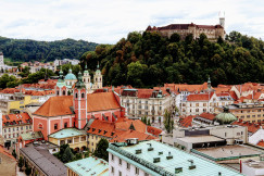 The photo presents a city with red-roofed buildings and church towers beneath a castle on a wooded hill.