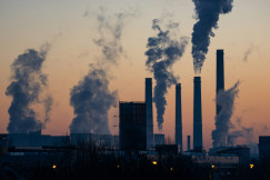 The photo presents an industrial facility with tall chimneys and large plumes rising into an orange sky.