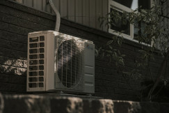 The photo presents an outdoor air-conditioning unit mounted beside a dark brick wall, with a leafy plant and window in the background.