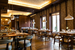 The photo presents an empty restaurant with set dining tables, wooden chairs, white pendant lights and large windows, surrounded by decorative wooden walls and warm lighting.