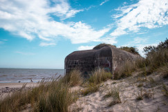 The photo presents a weathered concrete bunker among grassy sand dunes beside the sea, with two people and a dog walking along the beach.
