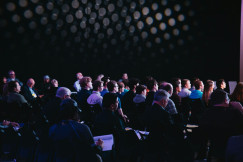 The photo presents an audience seated in rows in a dimly lit conference room, facing a presentation, with some attendees taking notes.