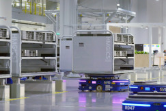 The photo presents mobile robots carrying large storage racks through a brightly lit industrial facility.