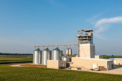 The photo presents a grain processing facility with tall metal silos and a large industrial tower surrounded by agricultural fields.