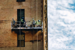 The picture shows the side of a tall brick building with two workers suspended on a motorized platform attached by vertical cables.