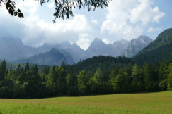 The photo presents a green meadow and dense forest beneath rocky mountain peaks partly covered by clouds.