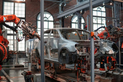 The photo presents orange industrial robotic arms surrounding an unfinished car body on an automotive assembly line.