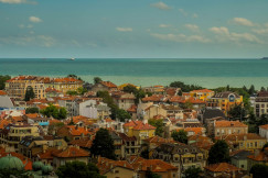 The photo presents a densely built coastal town with red-tiled rooftops overlooking the sea and several ships on the horizon.