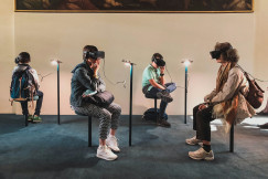 Four visitors wearing virtual reality headsets while seated in a museum or gallery space, with a large painting displayed on the wall above them.