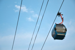 The photo presents a blue cable car suspended from overhead cables against a blue sky with a few white clouds.