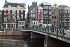 The photo presents people crossing a canal bridge lined with bicycles, with traditional canal houses in the background.