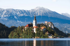 The photo presents a church on a wooded island in a calm lake, with a castle on a rocky cliff and forested mountains in the background.