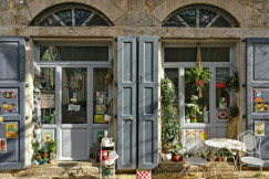 The photo presents a charming café storefront in a stone building, with grey shutters, potted plants, food posters and a small outdoor table with chairs.