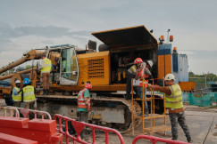 The picture describes a construction site where several workers wearing hard hats and high-visibility safety vests are inspecting and maintaining a large piece of heavy construction machinery.