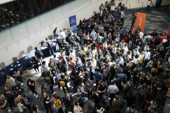 The photo presents an overhead view of people networking in a busy conference hall, with exhibition tables and banners around them.