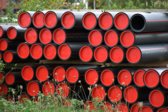 The photo presents stacks of large black industrial pipes with bright red protective caps, surrounded by vegetation.