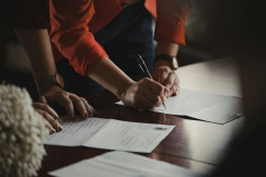 The photo presents a person signing a document at a wooden table, with other people and papers nearby.