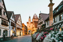 The photo presents a cobbled street lined with traditional timber-framed houses, flowers and a clock tower.