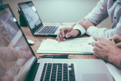 The photo presents two people reviewing handwritten notes and diagrams at a wooden table with two open laptops.