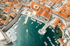 The photo presents an aerial view of a harbour filled with boats, surrounded by stone buildings with red-tiled roofs.
