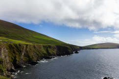The photo presents green hills and rocky cliffs along the sea under a partly cloudy sky.