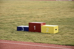 The photo presents an empty winners’ podium with red, yellow and blue blocks numbered one, two and three on a grassy field beside a running track.