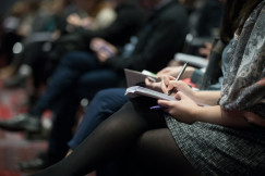 The photo presents a person taking notes in a notebook while seated among other attendees at an event.