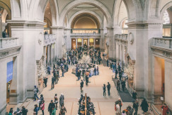 The photo presents a busy museum hall with ornate arches, columns, balconies and visitors gathered around exhibits and information desks.