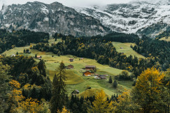 The photo presents scattered wooden houses on green hills, surrounded by forests and autumn foliage beneath snow-covered mountains.