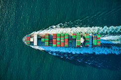 The photo presents an aerial view of a cargo ship carrying colourful containers through deep blue water, leaving a white wake behind.