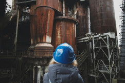 The photo presents a person wearing a blue hard hat facing large rusted industrial structures, metal walkways and staircases.