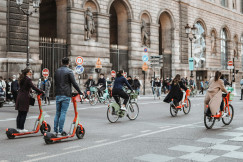 The picture shows people using bicycles and electric scooters in a busy urban street.