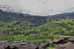 The photo presents a mountain village with wooden chalets scattered across green slopes, a cable car line and forested hills beneath snow-covered peaks.