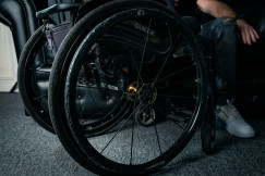 The photo presents a close-up of a wheelchair indoors, with its large wheels in the foreground and a seated person partially visible beside it.