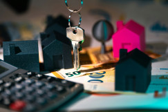The picture shows house-shaped models, a set of keys, euro banknotes, and a calculator, representing real estate, home ownership, and property financing.