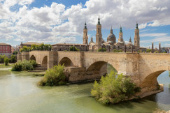 The photo presents a historic stone bridge and cathedral in Zaragoza, Spain.