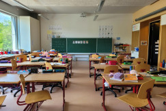 The picture shows a bright, well-organized classroom with rows of desks and chairs, school supplies, books, and a large chalkboard at the front.