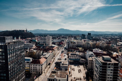 The picture shows an aerial view of an urban landscape in Slovenia, with a mix of modern and historic buildings and mountains in the background.