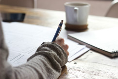 Colour photograph of a person writing on brown wooden table near white ceramic mug