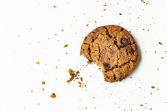 The photo presents a partially eaten chocolate chip cookie surrounded by crumbs on a white background.
