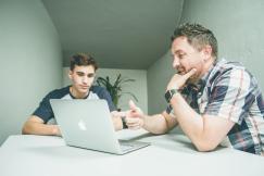 An illustrative photo of a man wearing white and black plaid button up sports shirt pointing the silver macbook