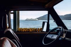 An illustrative photo of the interior of a vehicle with a steering wheel in the foreground and a body of water with a ferry in the distance.