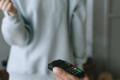 An illustrative photo of a person holding a phone with another person standing and holding a book in the background
