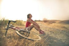 An illustrative photo of a sportive woman with a bicycle resting on a countryside road in sunlight