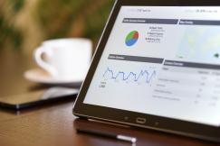 An illustrative photo of a tablet displaying analytics charts and graphs, with a smartphone and a cup of coffee in the background